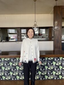 Chieko.M standing and smiling in a bright café-style classroom, wearing a white cardigan with colorful embroidery
