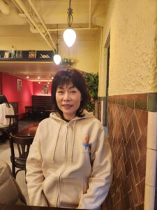 Tomoko.T, seated in a cozy café, smiling in a beige hoodie